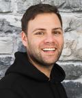 Florian Auf dem Bild sieht man einen jungen Mann, der einen schwarzen Pulli trägt und freundlich in die Kamera schaut. Der Hintergrund zeigt eine Steinwand.