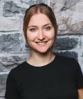 Auf dem Bild sieht man eine junge Frau mit dunkelblonden Haaren und einem schwarzen T-Shirt, die vor einer Steinwand steht und freundlich in die Kamera schaut.