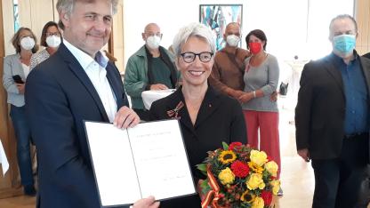 Auf dem Bild sieht man den Karlsruher OB und Karin, die beide lächelnd in die Kamera schauen. Karin trägt das Bundesverdienstkreuz und hält einen Blumenstrauß in der Hand.  